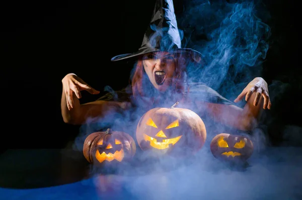 An ominous witch in a hat conjures over a jack-o-lantern. Traditional halloween characters. Mystical fog creeps over pumpkins with carved terrible faces. Woman in carnival costume.