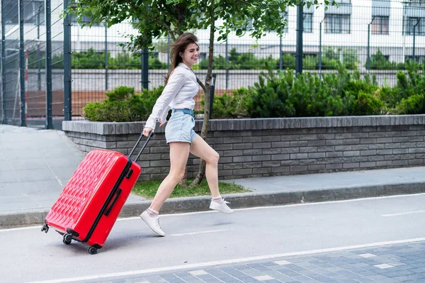 Glücklich schöne kaukasische Frau läuft mit einem großen roten Koffer nach draußen. Aufgeregtes Mädchen kommt zu spät ins Flugzeug. — Stockfoto