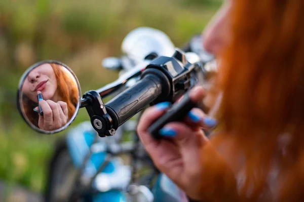 Kobieta na motocyklu patrzy w lustro i maluje usta szminką. Refleksja rudej rowerzystki. — Zdjęcie stockowe