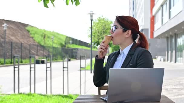 Uśmiechnięta kobieta pracująca zdalnie na laptopie i jedząca lody siedząc przy drewnianym stole na świeżym powietrzu. Portret dziewczyny w garniturze służbowym na przerwę obiadową w ulicznej kawiarni. — Wideo stockowe