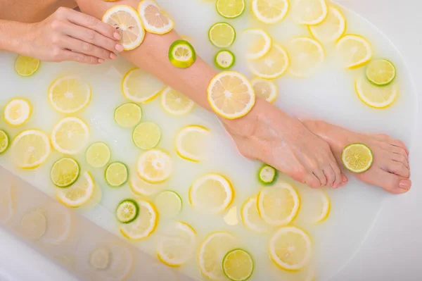 Mujer irreconocible toma un baño de leche con limones y limas. Spa cítrico. Cuidado corporal. Blanqueamiento de la piel. piernas femeninas vista superior. — Foto de Stock