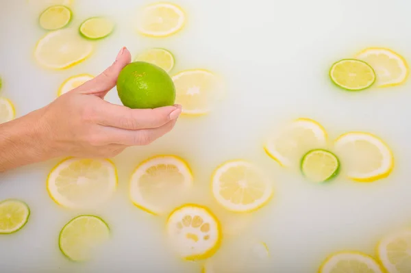 Gros plan d'une main féminine tenant un citron vert sur de l'eau blanche avec des tranches de citron — Photo