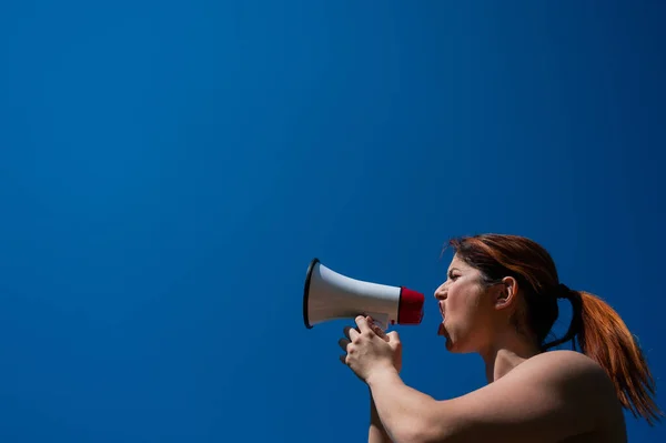 Рыжая белая женщина кричит в мегафон на фоне голубого неба. Девушка делает объявление через громкоговоритель. — стоковое фото