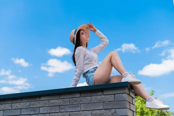 Mulher bonita em shorts e um chapéu posando enquanto sentado em um banco de pedra. Um retrato completo de uma menina caucasiana em um dia quente de verão. — Fotografia de Stock