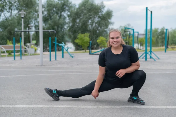 Fettleibige junge Kaukasierin beim Seitensprung. Dicke schöne lächelnde Mädchen in einem schwarzen Trainingsanzug ist in Fitness im Freien beschäftigt. Aufwärmen vor dem Training. — Stockfoto