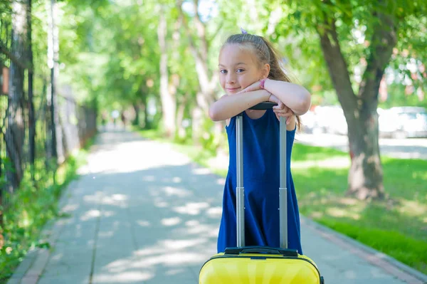Spokojená holčička v modrých šatech stojí sama na ulici se žlutým kufříkem a čeká na rodiče. Dítě jede na letní prázdninový výlet. — Stock fotografie