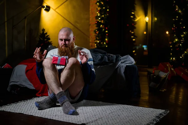 Egy hosszú, vörös szakállú férfi ül a földön nadrág nélkül egy téli pulóverben. Az ember egy becsomagolt ajándékot tart az újévi dekorációk és fények hátterében. Karácsonyfa. Paródia, humor. — Stock Fotó