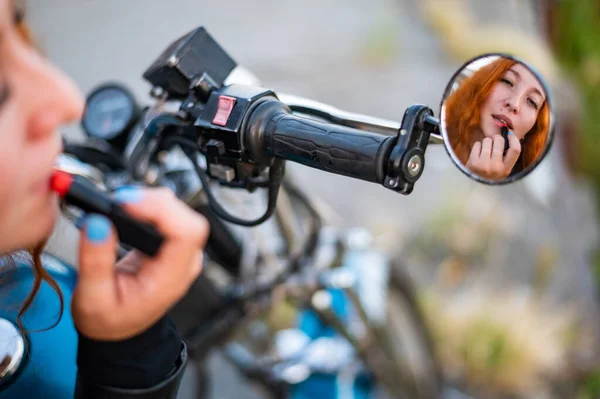 骑摩托车的女人照镜子，涂口红。一个红头发的骑自行车的女孩的倒影. — 图库照片