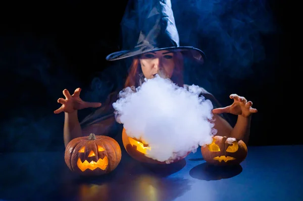 An ominous witch in a hat conjures over a jack-o-lantern. Traditional halloween characters. Mystical fog creeps over pumpkins with carved terrible faces. An evil witch casts a spell. — Stock Photo, Image