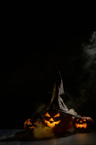 Κάθετη κάρτα Halloween. Καπέλο μάγισσας σε κολοκύθα με σκαλιστά ανατριχιαστικά γκριμάτσες σε μαύρο φόντο στην ομίχλη. Ο Jack-o-lantern λάμπει από μέσα και εκπέμπει καπνό. — Φωτογραφία Αρχείου