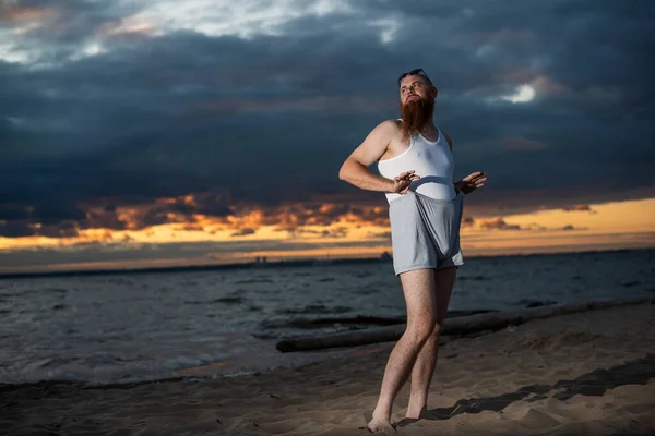 Een brutale kale man poseert op het strand bij zonsondergang parodie glamoureuze chick — Stockfoto