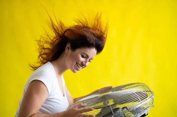 Una hermosa mujer pelirroja se enfría de pie sobre un gran ventilador eléctrico sobre un fondo amarillo. Chica con el pelo en desarrollo en el viento sobre Dispositivo para enfriar el aire — Foto de Stock