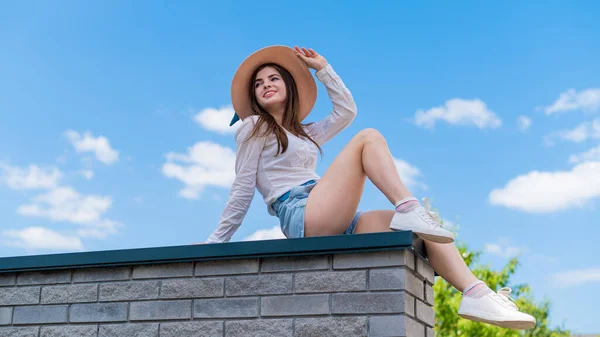 Vacker ung kvinna i shorts och en hatt poserar sittande på en stenbänk. Ett långt porträtt av en vit flicka en varm sommardag.. — Stockfoto