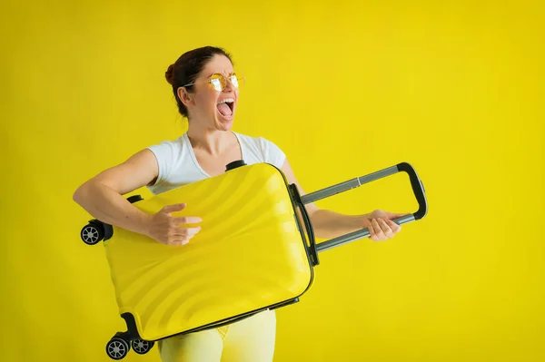 Vacker kaukasisk kvinna lurar runt med en resväska på en gul bakgrund. En charmig tjej imiterar att hon spelar gitarr. Glädjen i den kommande resan. — Stockfoto