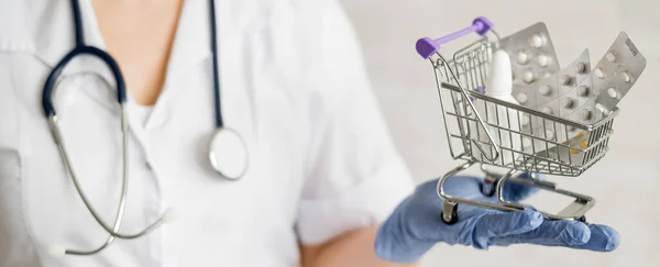 Um médico sem rosto com um casaco médico e um estetoscópio segura um pequeno carrinho de compras cheio de medicamentos. Conceito de farmácia. — Fotografia de Stock