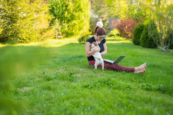 Mulher caucasiana está digitando em um laptop enquanto está sentado em um gramado verde ao ar livre. Freelancer feminino trabalha remotamente no parque . — Fotografia de Stock