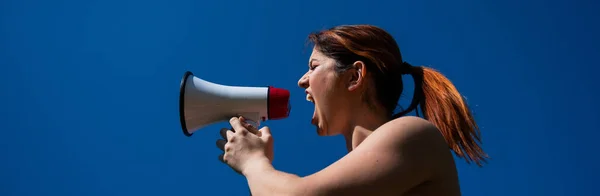 Рыжая белая женщина кричит в мегафон на фоне голубого неба. Девушка делает объявление через громкоговоритель. — стоковое фото