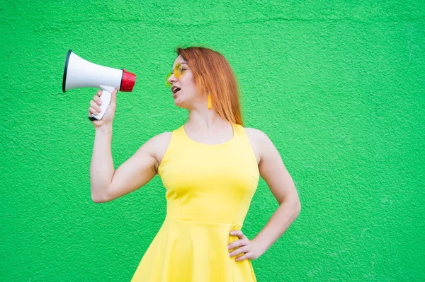 Rothaarige Frau mit Sonnenbrille und gelbem Kleid hält ein Megafon auf grünem Hintergrund. — Stockfoto