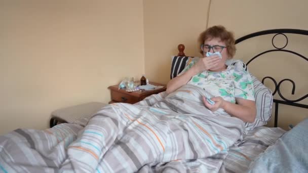 Egy influenzás idős nő fekszik az ágyban. A nyugdíjas tüsszent és belefújja az orrát egy szalvétába.. — Stock videók