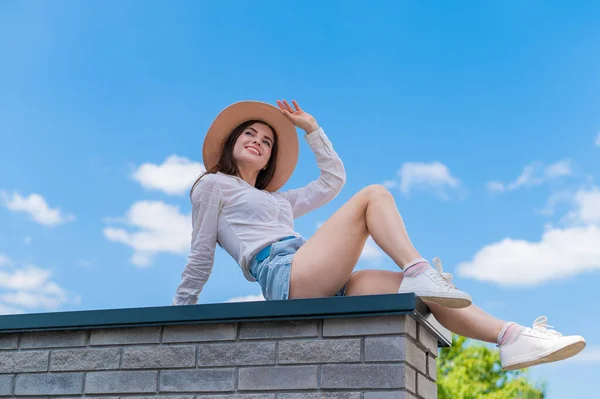 Hermosa joven en pantalones cortos y un sombrero posando mientras está sentada en un banco de piedra. Un retrato completo de una chica caucásica en un caluroso día de verano. —  Fotos de Stock