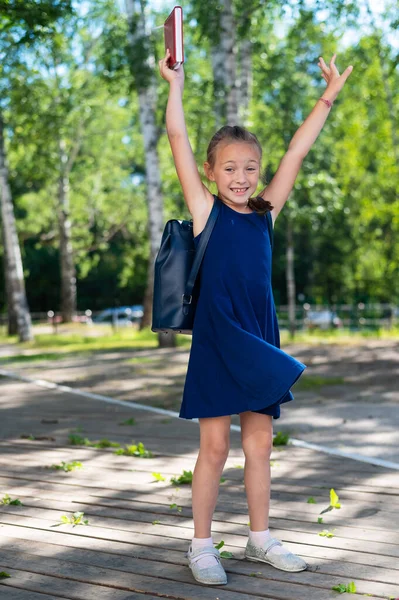 Opgewonden schoolmeisje houdt een leerboek vast en springt in het park. Het meisje is blij om terug naar school te gaan.. — Stockfoto