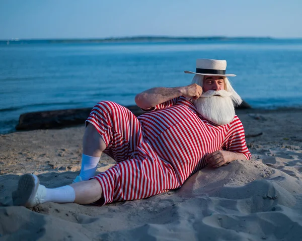 Un uomo anziano in costume da bagno retrò a righe prende il sole sulla spiaggia. Un vecchio uomo barbuto dai capelli grigi con un cappello giace sulla sabbia vicino al mare. — Foto Stock