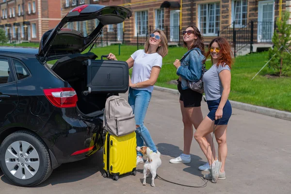 Drei Freundinnen gehen auf Roadtrip. Frauen laden ihr Gepäck in den Kofferraum eines Autos. Mädchen fahren mit dem Hund in den Urlaub. — Stockfoto