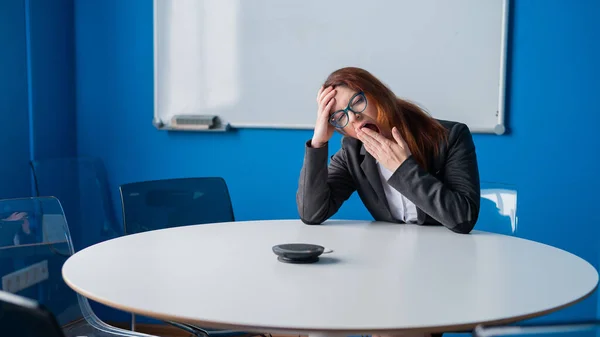 Trött rödhårig affärskvinna i glasögon och en kostym uttråkad i ett tomt konferensrum. En kontorist gäspar väntar på början av en presentation i styrelserummet. — Stockfoto