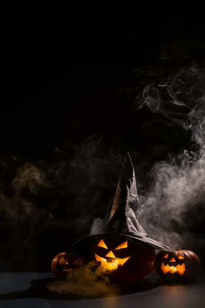 Tarjeta vertical de Halloween. Sombrero de bruja en una calabaza con muecas espeluznantes talladas sobre un fondo negro en la niebla. Jack-o-linterna brilla desde el interior y emite humo. — Foto de Stock