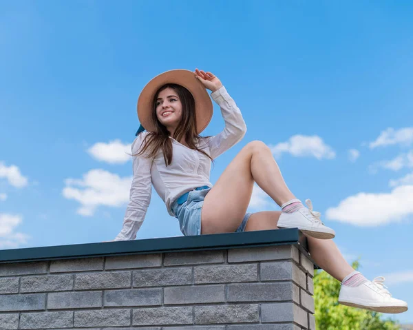 穿着短裤,头戴帽子的年轻漂亮女子坐在石凳上摆姿势.一个白种人女孩在炎热的夏天的全长肖像. — 图库照片