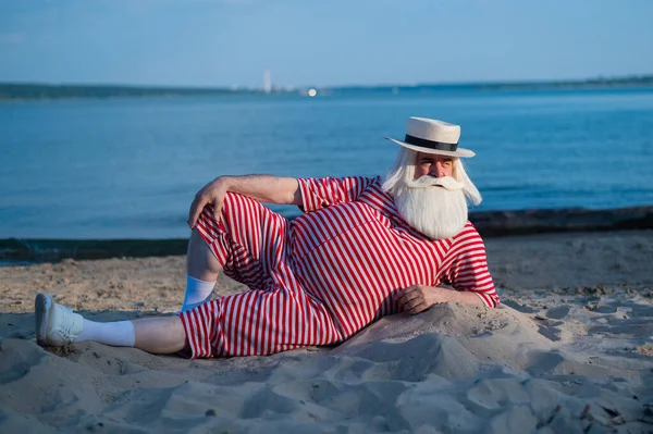 Un uomo anziano in costume da bagno retrò a righe prende il sole sulla spiaggia. Un vecchio uomo barbuto dai capelli grigi con un cappello giace sulla sabbia vicino al mare. — Foto Stock