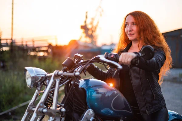 Mulher ruiva encaracolado em uma jaqueta de couro preto senta-se em uma motocicleta ao pôr do sol. Retrato de uma menina séria dirigindo uma bicicleta. — Fotografia de Stock