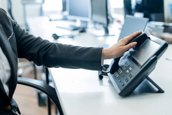 Κοντινό γυναικείο χέρι στο σταθερό τηλέφωνο στο γραφείο. Ανέκφραστη γυναίκα με κοστούμι εργάζεται ως ρεσεψιονίστ απαντώντας στο τηλέφωνο σε κλήσεις πελατών. — Φωτογραφία Αρχείου