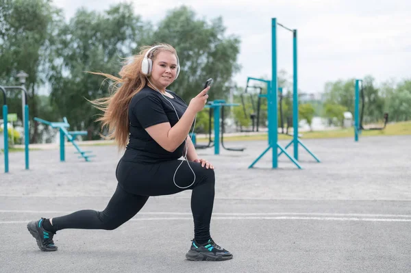 Uma linda garota gorda em fones de ouvido está envolvida na aptidão no campo esportivo e tira uma selfie. Jovem mulher lunges e é fotografado em um smartphone ao ar livre em um dia quente de verão. — Fotografia de Stock