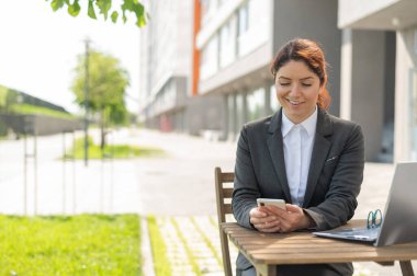 Gülümseyen beyaz kadın yaz terasında ahşap bir masada oturuyor ve akıllı telefon kullanıyor.