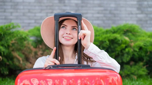 Młoda, piękna, biała kobieta w kapeluszu wygląda z czerwonej walizki z rączką. Szczęśliwa dziewczyna jedzie na wycieczkę z bagażem. Koncepcja wakacji letnich. — Zdjęcie stockowe
