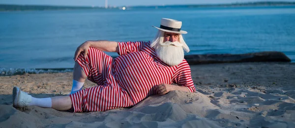 Пожилой мужчина в полосатом ретро купальнике загорает на пляже. Старый седой бородатый мужчина в шляпе лежит на песке у моря. — стоковое фото