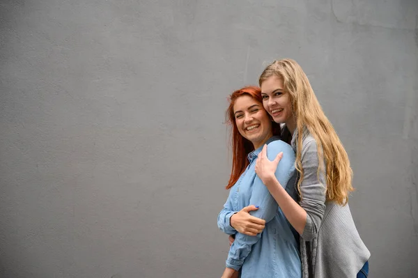 Twee blije vriendinnen knuffelen op de achtergrond van een grijze muur. Zachte knuffels van een lesbisch echtpaar. LGBT huwelijk van hetzelfde geslacht. — Stockfoto