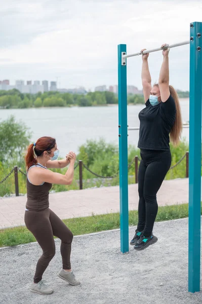 A female personal trainer helps a fat client to pull herself up on horizontal bars correctly. Two women wearing protective masks training outdoors during the coronavirus outbreak.