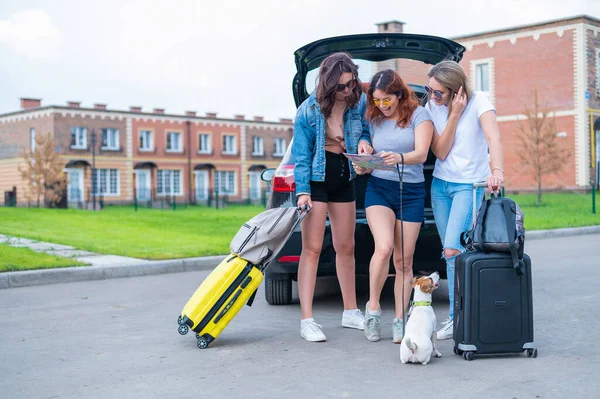 三个朋友正在路上旅行过暑假，正在看地图。年轻妇女把手提箱装进汽车后备箱. — 图库照片