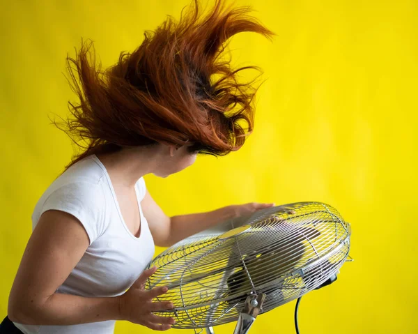 Una hermosa mujer pelirroja se enfría de pie sobre un gran ventilador eléctrico sobre un fondo amarillo — Foto de Stock