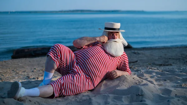 Un anciano con un traje de baño retro a rayas toma el sol en la playa. Un viejo barbudo de pelo gris con sombrero yace en la arena junto al mar. — Foto de Stock