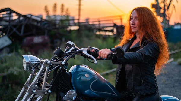 Mulher ruiva encaracolado em uma jaqueta de couro preto senta-se em uma motocicleta ao pôr do sol. Retrato de uma menina séria dirigindo uma bicicleta. — Fotografia de Stock