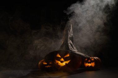 Sisin ortasında karanlıkta üç tane balkabağı feneri parlıyor. Cadılar Bayramı kabağı ve cadı şapkası..