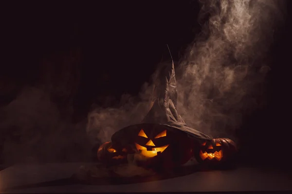 霧の中、暗闇の中で3つのジャック・オ・ランタンが輝きます。魔女の帽子のハロウィンカボチャ. — ストック写真