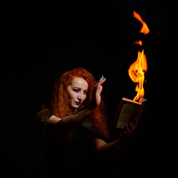 An ominous witch sets her spell book on fire with the power of thought. Red-haired woman conjures for Halloween. Flames on the hands.