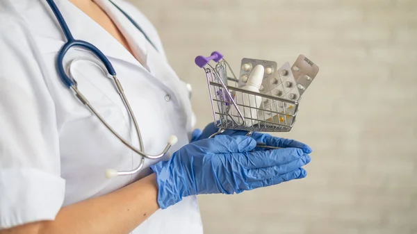 Egy arctalan orvos orvosi kabáttal és sztetoszkóppal tart egy kis bevásárlókocsit tele gyógyszerekkel. Gyógyszerészeti fogalom. — Stock Fotó