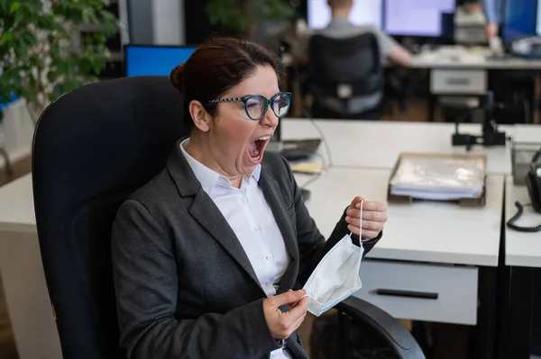 En kvinne i dress sitter på en arbeidsplass og gjesper mens hun tar av seg masken.. – stockfoto