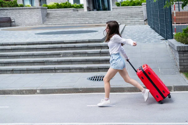 Glücklich schöne kaukasische Frau läuft mit einem großen roten Koffer nach draußen. Aufgeregtes Mädchen kommt zu spät ins Flugzeug. — Stockfoto