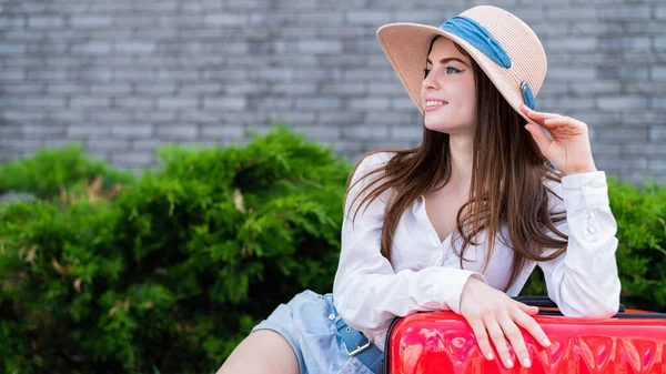 Portret van een mooie blanke vrouw in een hoed met een grote rode koffer buiten. Aantrekkelijk meisje leunde tegen bagage tijdens het wachten op een taxi. — Stockfoto